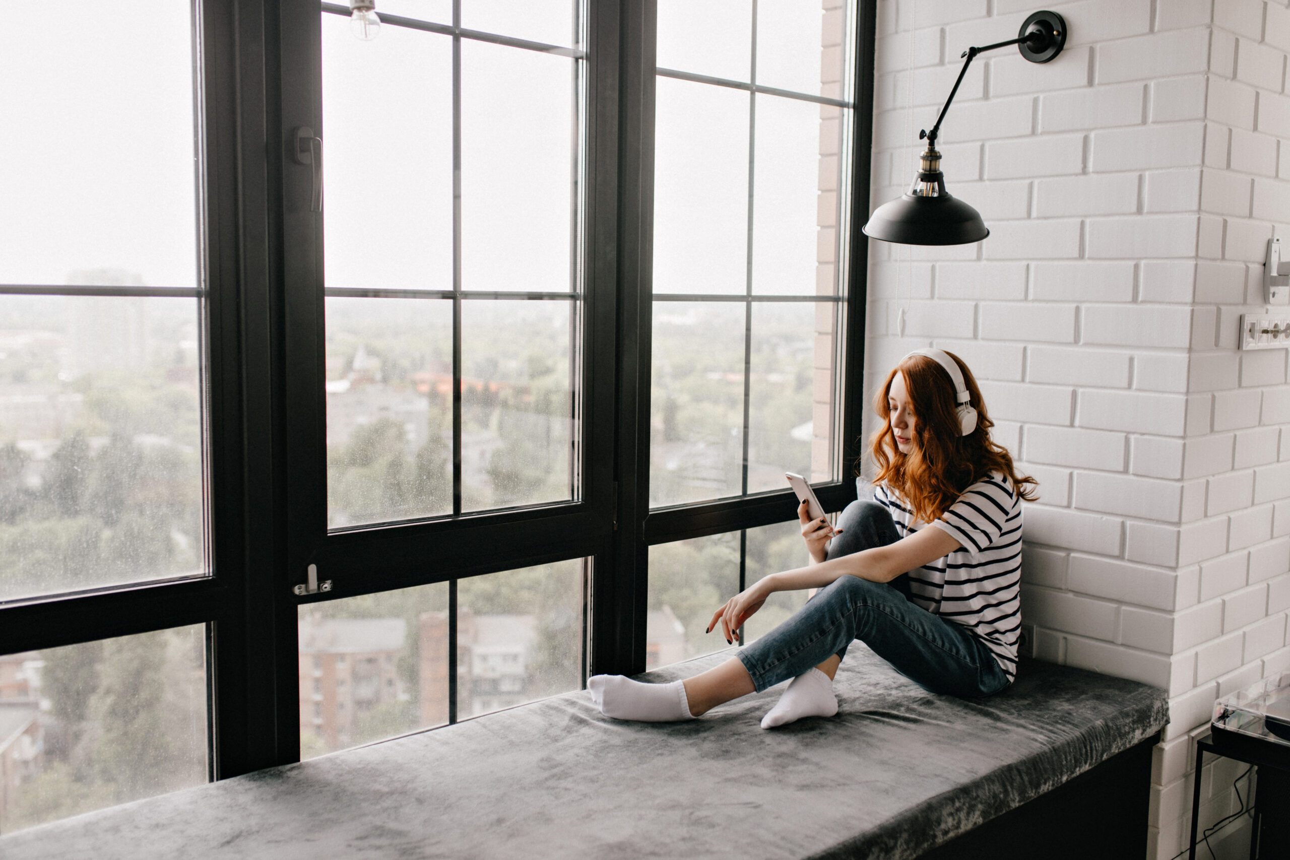 Adorable red-haired girl in headphones sitting on window sill. I Adorable red-haired girl in headphones sitting on window sill. Indoor shot of pensive ginger lady using phone at home.